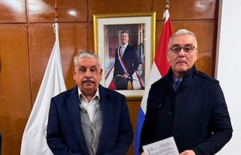 Isaías Fretes con chaleco y Robert Rivarola en abrigo oscuro, posando en oficina con cuadro y banderas de fondo.