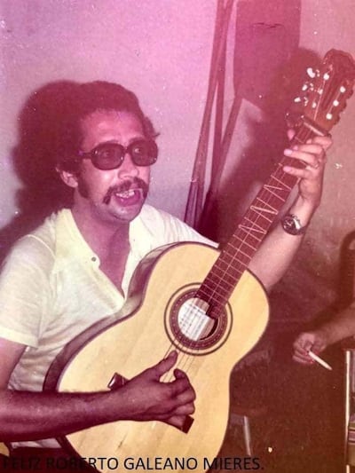 Maneco Galeano, con gafas oscuras y camisa amarilla, toca la guitarra mientras mira a un público atento.