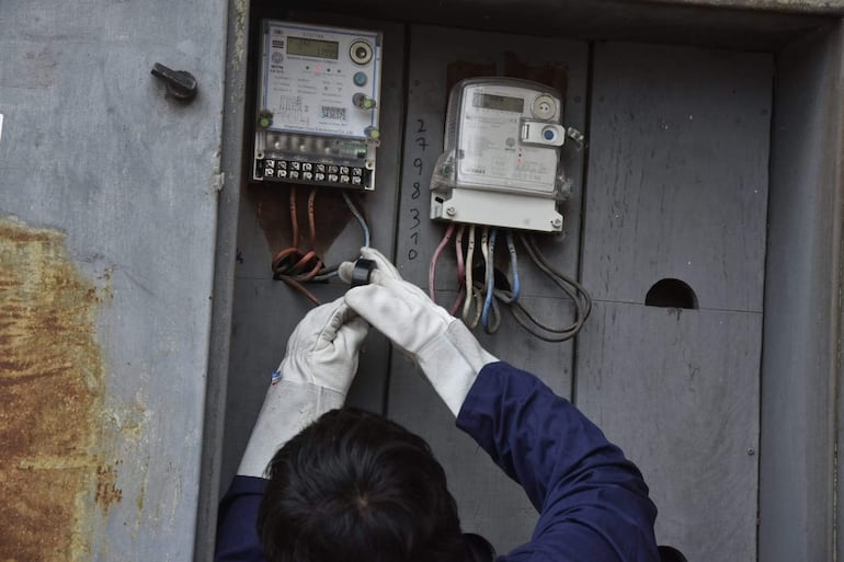 Funcionario en guantes blancos y chaqueta oscura trabaja en panel eléctrico con medidores desconectados en un entorno exterior.