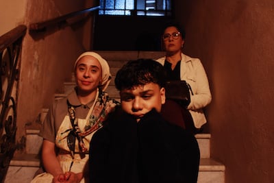 Mujer con vestido claro y pañuelo sonríe, joven pensativo en el centro y mujer mayor con gafas observa a la derecha en una escalera.