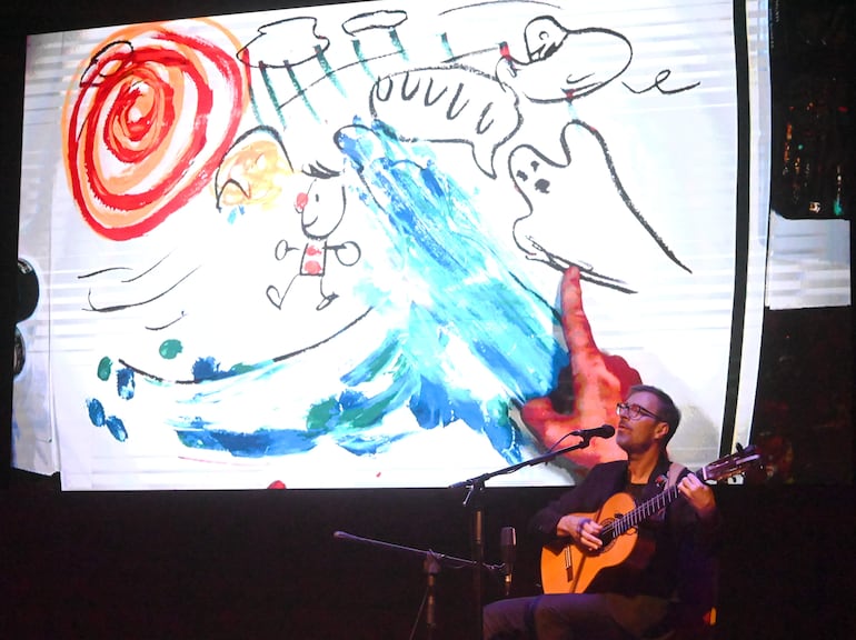 Kevin Johansen, con gafas y camisa oscura, toca guitarra en el escenario mientras se proyecta arte colorido detrás.