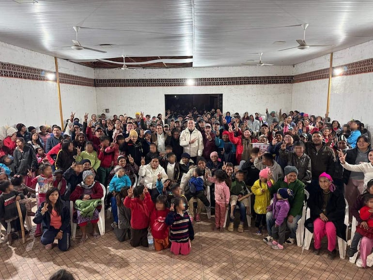 Cientos de personas levantando las manos en señal de paz, con atuendos coloridos en un salón con paredes de cemento.