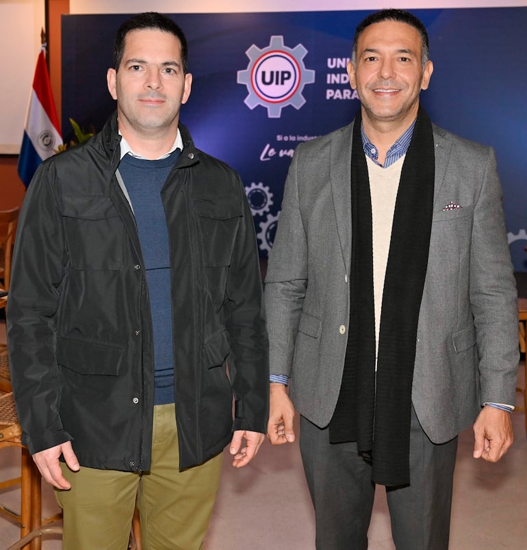 Dos hombres en chaquetas, sonriendo en un evento de la UIP, con el logo y la bandera de Paraguay al fondo.