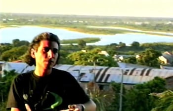 Feliciano Centurión sentado, gesticulando con camiseta oscura y cabello largo en un paisaje natural junto al río.