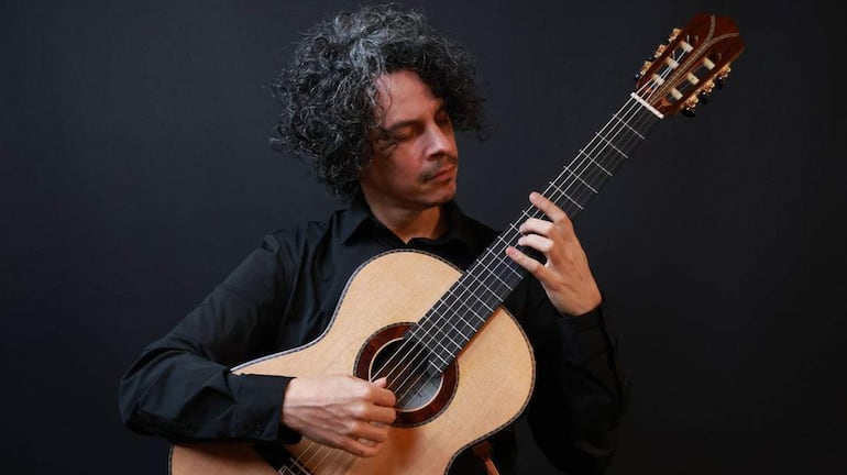 Alí Arango, con cabello rizado y camisa negra, toca guitarra en un fondo negro, mostrando concentración en su actuación.