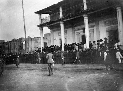 Multitud expectante en exterior; grupo formal en balcón y soldado al centro, observando atentamente el evento.