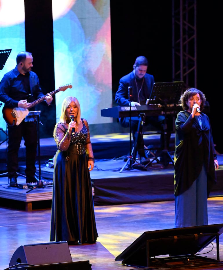 Cantantes de Mocedades en el escenario, una en vestido brillante y otra en ropaje oscuro, rodeadas de músicos y luces de colores.