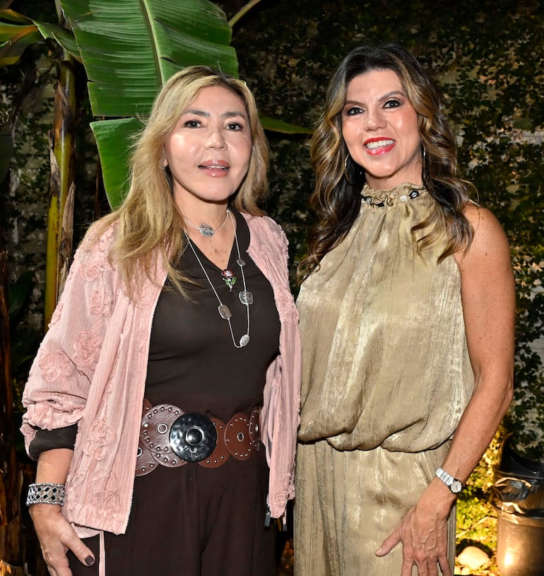 Dos mujeres sonrientes, una en chaqueta rosa y otra en vestido dorado, rodeadas de vegetación en un evento nocturno.
