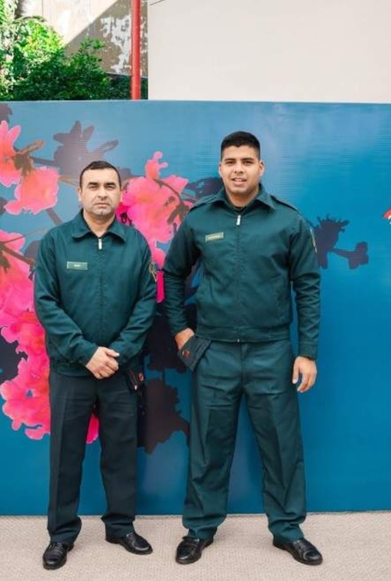 Dos hombres en uniformes oscuros, uno con expresión seria y el otro sonriendo, posan en un fondo decorativo colorido.