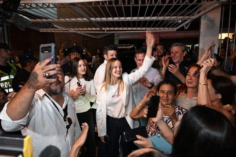 Soledad Núñez sonriente con blazer blanco en el centro, rodeada de personas aplaudiendo y policías de fondo.