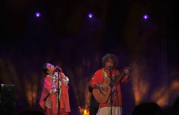 Jennifer Hicks canta con micrófono y capa roja, mientras Miguel Narváez toca la guitarra; ambiente de concierto con iluminación teatral.
