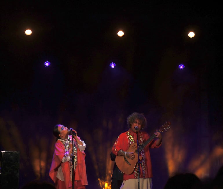 Jennifer Hicks canta con micrófono y capa roja, mientras Miguel Narváez toca la guitarra; ambiente de concierto con iluminación teatral.