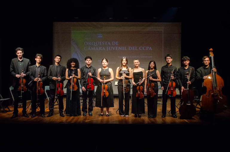 Doce jóvenes músicos de la Orquesta de Cámara Juvenil del CCPA, vestidos formalmente, listos para tocar, con instrumentos de cuerda en mano.
