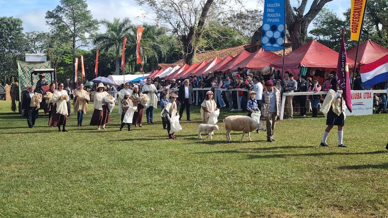 Grupo de 15 personas en trajes tradicionales caminan con un cordero entre ellos, rodeados de una multitud en un festivo ambiente al aire libre.