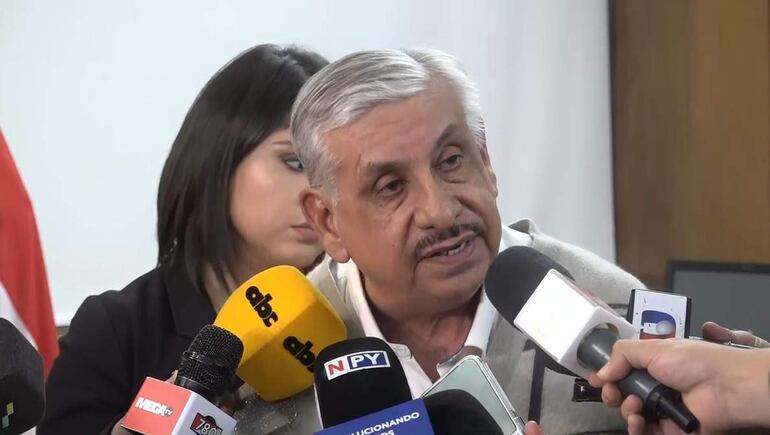 Hombre canoso con camisa clara y suéter, habla ante micrófonos; mujer de cabello largo observa en segundo plano.