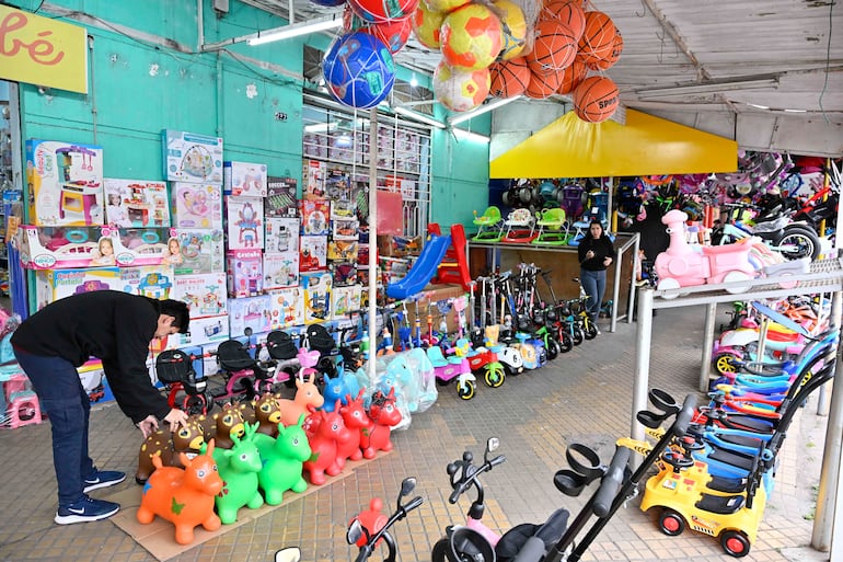 Hombre agachado en un mercado de juguetes, rodeado de mujeres examinando productos coloridos para niños.