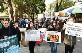 Dos mujeres sostienen carteles de protesta en la plaza, una con cabello oscuro y otra con abrigo negro, rodeadas de otros manifestantes.
