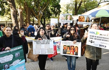 Dos mujeres sostienen carteles de protesta en la plaza, una con cabello oscuro y otra con abrigo negro, rodeadas de otros manifestantes.