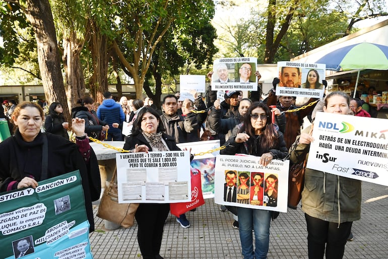 Dos mujeres sostienen carteles de protesta en la plaza, una con cabello oscuro y otra con abrigo negro, rodeadas de otros manifestantes.