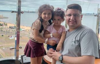 Hombre de gafas con una niña en brazos y otra a su lado, sonriendo, en un área recreativa con fondo urbano.