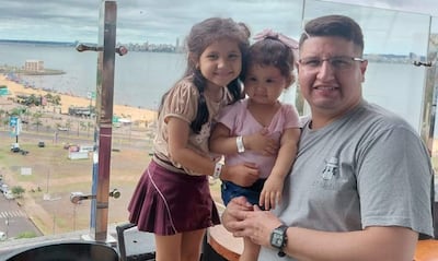 Hombre de gafas con una niña en brazos y otra a su lado, sonriendo, en un área recreativa con fondo urbano.