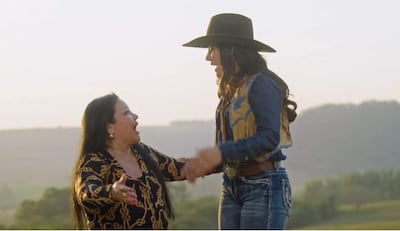 Perla gesticula con manos, vestida con blusa oscura y cabello largo. Fiorella luce camisa de mezclilla y sombrero negro, en un campo abierto.