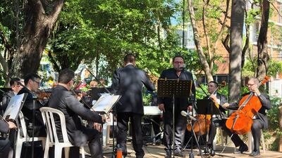 Grupo de músicos en trajes oscuros, con un cantante al micrófono y un pianista en teclado, rodeados de un entorno natural.