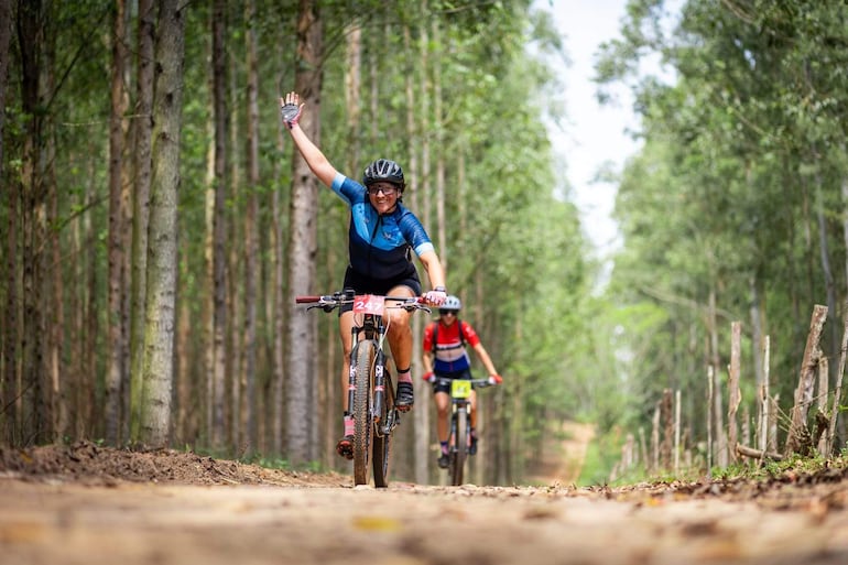 Mujer con camiseta azul saluda mientras ciclista con camiseta roja la sigue en un bosque, ambos con cascos y guantes.