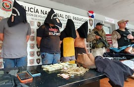 Cinco hombres con capuchas alineados frente a un oficial militar en un recinto policial, con billetes y cartuchos sobre la mesa.
