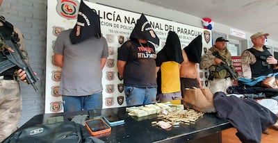 Cinco hombres con capuchas alineados frente a un oficial militar en un recinto policial, con billetes y cartuchos sobre la mesa.