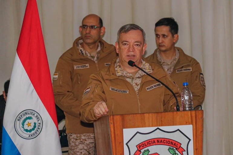 César Roberto Silguero Lobos habla en podio con chaqueta marrón claro, rodeado de otros tres hombres en un auditorio con bandera paraguaya.