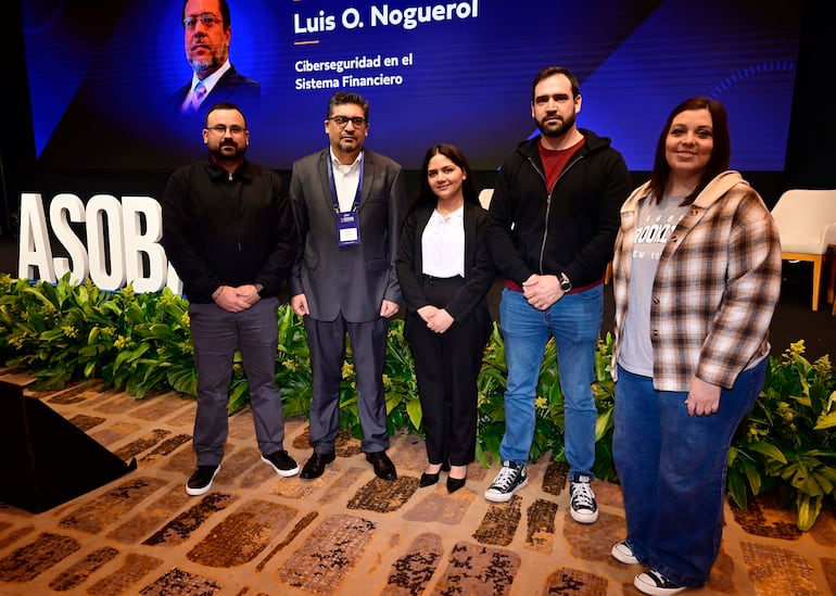 Luis O. Noguerol en traje negro presenta en panel, rodeado de cuatro personas, en auditorio iluminado con plantas decorativas.