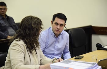 Hernán Rivas en chaqueta clara, sentado junto a un hombre de camisa de cuadros. Mesas con documentos y un teléfono móvil están visibles.