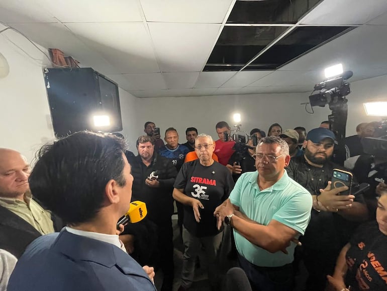 Julio Fernández en traje oscuro, hablando ante un grupo atento; el ambiente es iluminado y se ven teléfonos y cámaras.