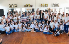 Treinta personas en camisetas blancas sosteniendo retratos y trofeos en salón festivo, con niños en primer plano mostrando orgullo.