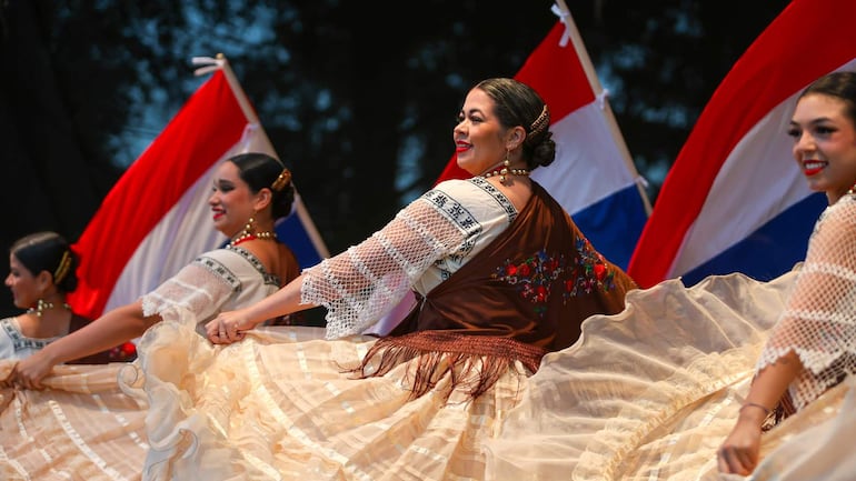 Cuatro bailarinas en trajes tradicionales sonríen mientras realizan un baile folklórico sobre un escenario oscurecido con banderas paraguayas de fondo.