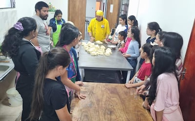 Niños y un hombre con delantal amarillo en cocina, observando una preparación en mesa de acero con masa para chipitas.