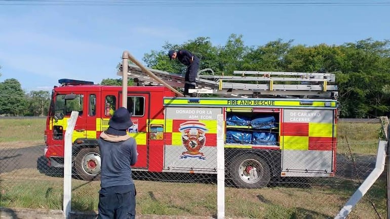 Bombero en camión rojo con franjas amarillas, mientras un observador de espaldas observa la acción en un paisaje verde.
