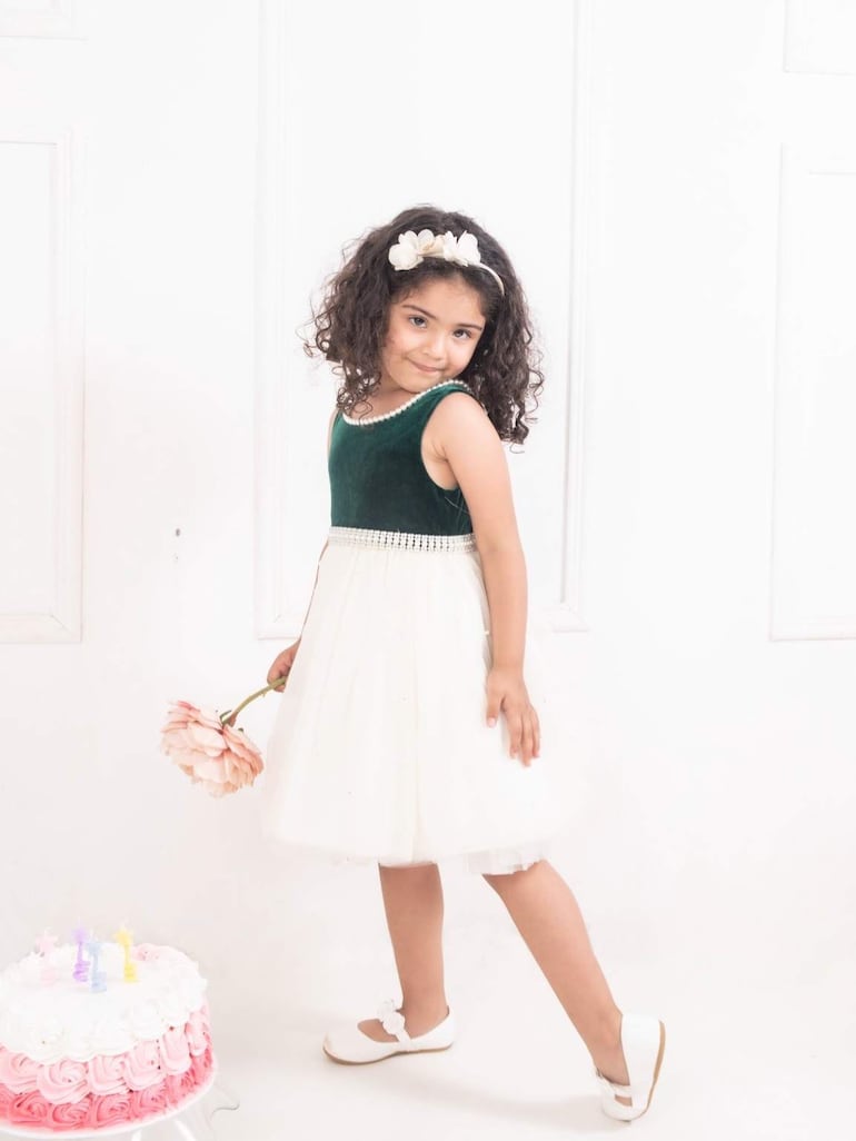 Niña con cabello rizado y vestido blanco sostiene una flor, junto a una torta rosa y blanca con velas encendidas.