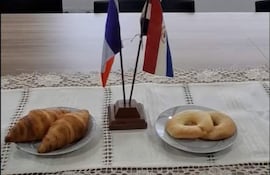 Mesa con mantel de encaje, tres croissants a la izquierda y chipa a la derecha, decorada con banderas de Paraguay y Francia.