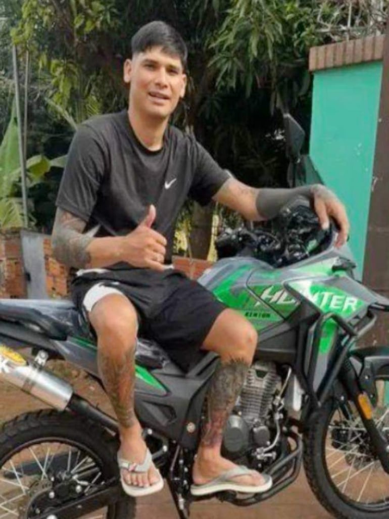 Verón, con camiseta negra y short, sonríe sentado en motocicleta, gesticulando un pulgar arriba en ambiente exterior.