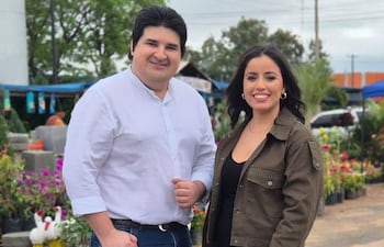 Montefilpo con camisa blanca y jeans, Lezcano en blusa negra y chaqueta verde, sonrientes en un colorido mercado.