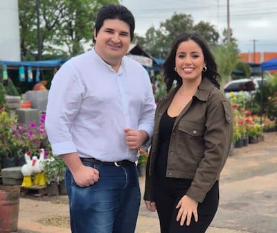 Montefilpo con camisa blanca y jeans, Lezcano en blusa negra y chaqueta verde, sonrientes en un colorido mercado.