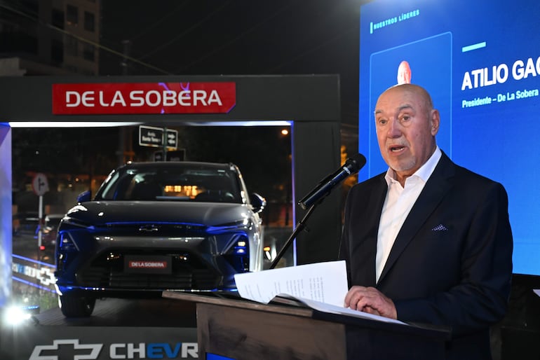 Hombre mayor, Atilio Ga De La Sobera, vestido formalmente, habla frente a un micrófono en un evento nocturno sobre automóviles Chevrolet.
