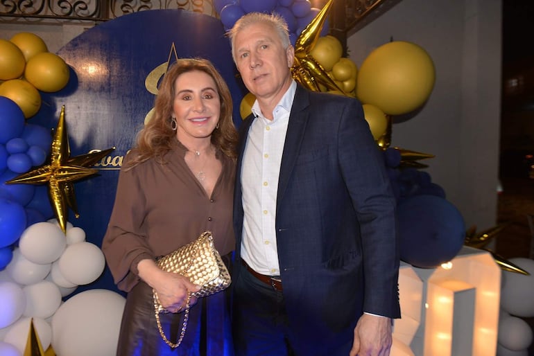 Mujer con blusa marrón y bolso dorado sonríe junto a hombre con camisa blanca y saco oscuro. Ambiente festivo decorado con globos.