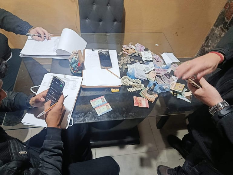 Tres personas en interior: dos sentadas revisan documentos y dinero, una de pie cuenta efectivo, rodeados de dispositivos móviles.