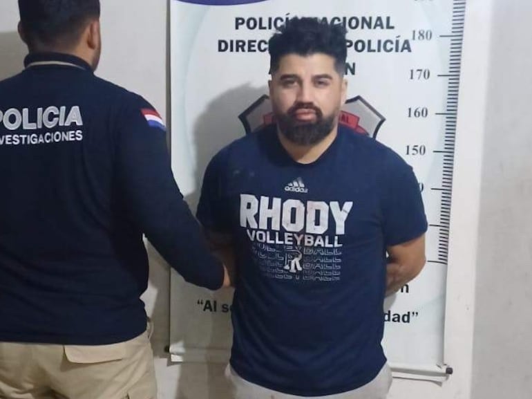 Hombre detenido con camiseta oscura en los brazos cruzados, mientras un oficial de policía de espaldas observa en oficina sencilla.