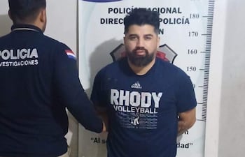 Hombre detenido con camiseta oscura en los brazos cruzados, mientras un oficial de policía de espaldas observa en oficina sencilla.