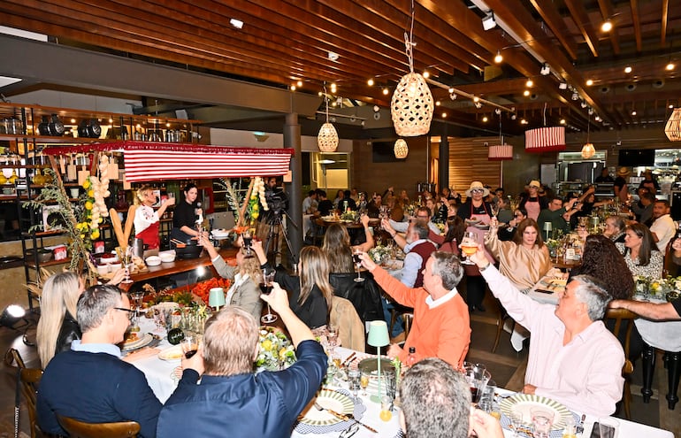 Grupo de 30 personas brindando en mesa elegante, cocina al fondo con mujer de delantal rojo y otra asistiendo en preparación.