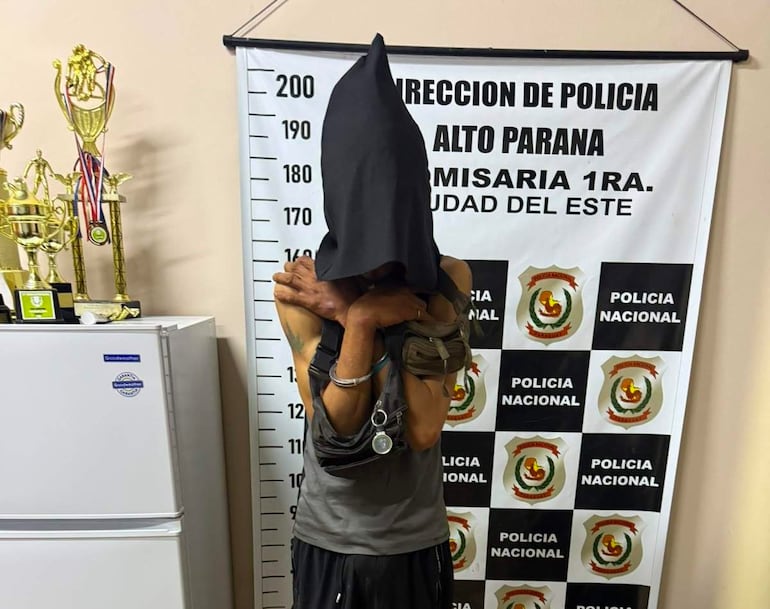 Hombre con capucha negra y grilletes en postura encorvada, con cartel de policía de fondo en comisaría.